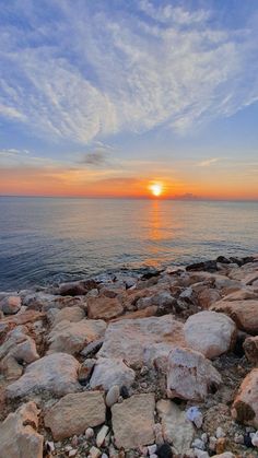 the sun is setting over the ocean with rocks and pebbles on the ground in front of it