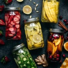 several jars filled with different types of fruit and veggies next to sliced lemons, strawberries, apples, oranges, cranberries