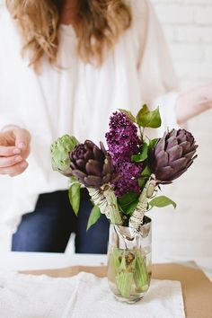 a woman sitting at a table with a vase filled with purple flowers and greenery