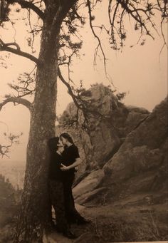 an old black and white photo of two people hugging under a tree in the mountains