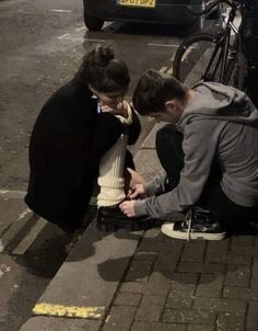 two people sitting on the sidewalk next to each other looking at something in their hands