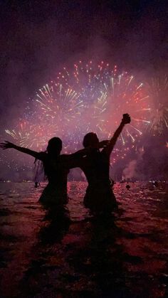 two people standing in the water with their arms around each other as fireworks go off behind them