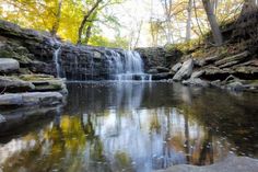 a small waterfall in the middle of a forest filled with trees and rocks, surrounded by fall foliage