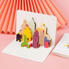 a pop up card with city buildings on it next to a folded book and paper fan