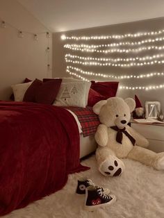 a teddy bear sitting on the floor in front of a bed with red sheets and lights