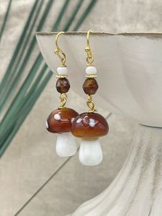 Handmade, adorable lampwork glass mushroom earrings. These earrings feature a charming brown glass mushroom bead with delicate white spots. The brown glass mushroom bead is skillfully paired with gold-plated wires and complemented by matching brown Czech glass beads. These trendy mushroom earrings are perfect for adding a touch of whimsy to your everyday outfits.  They would make the perfect gift for nature enthusiasts or anyone who loves unique accessories. For your convenience and peace of min Glass Mushroom Earrings, Mushroom Bead Earrings, White Mushroom Design Drop Earrings, White Dangle Earrings With Mushroom Design, White Mushroom Design Earrings For Gift, White Earrings With Mushroom Design As Gift, White Mushroom Design Earrings As Gift, Whimsical White Mushroom Design Earrings, White Mushroom-shaped Jewelry With Mushroom Design