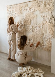 two women are working on a wall hanging with white yarn and balls in front of them