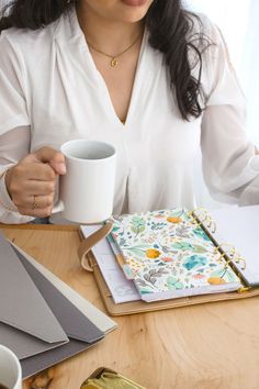 a woman sitting at a table holding a coffee cup and looking down on her notebook