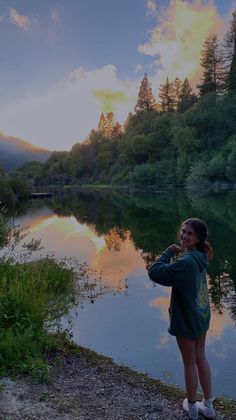 a woman standing next to a body of water with trees in the background at sunset