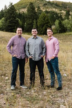 three men standing next to each other in a field with trees on the hill behind them