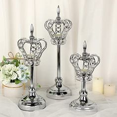 three silver candlesticks on a white table cloth with flowers and candle holders in the background