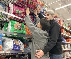 two men shopping in a grocery store with bags of cat food on the shelf and another man reaching for something