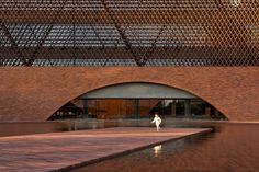 a person walking in front of a building with a large brick wall and water feature