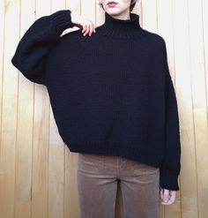 a woman standing in front of a wooden wall wearing a black sweater and brown pants