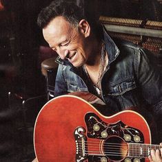 a man holding an acoustic guitar in his right hand and smiling at the camera while sitting down