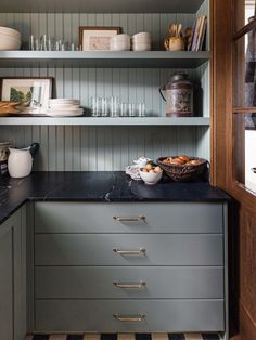 a kitchen with gray cabinets and black counter tops, white dishes on the shelves above