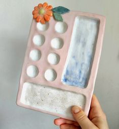 a hand holding up a pink and white ceramic object with flowers on the top, in front of a gray wall