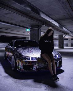 a woman sitting on the hood of a car in an underground parking garage with her skateboard