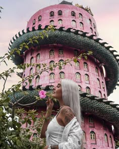 a woman with white hair standing in front of a pink building and holding a flower