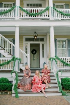 three women sitting on the front steps of a house decorated with christmas garland and lights