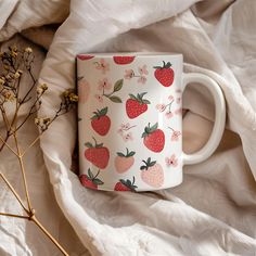 a coffee mug with strawberries on it sitting on a white blanket next to dried flowers