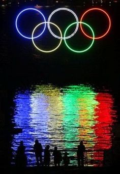 the olympic rings are lit up at night over water with people standing in front of it