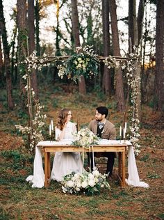 a man and woman sitting at a table in the woods with white flowers on it