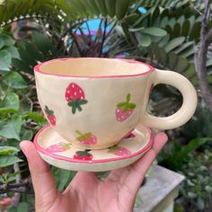 a hand holding a cup and saucer with strawberries on it in front of plants