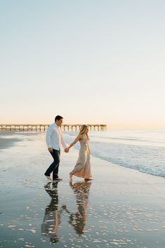 a man and woman holding hands walking on the beach at sunset with pier in background