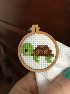 a hand holding a small cross stitch turtle on a white cloth with a wooden frame