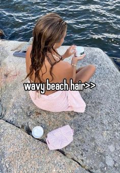 a woman sitting on top of a rock next to the ocean drinking from a cup