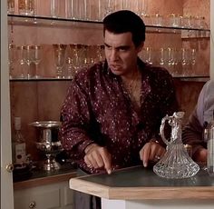 two men standing in front of a counter with wine glasses on it and one holding a glass pitcher