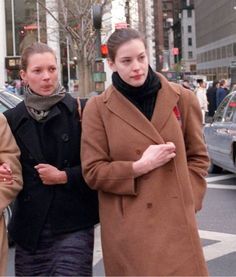 two women are walking down the street with their hands in their pockets and one is wearing a brown coat