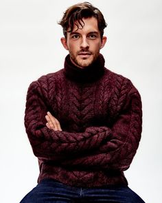 a man with his arms crossed wearing a turtle neck sweater and jeans, sitting on a stool