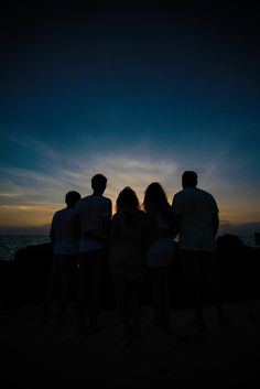 group of people standing on the beach at sunset with their arms around each other looking out to sea