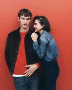 a man and woman standing next to each other in front of a red wall together