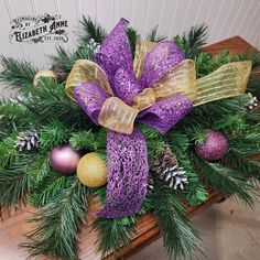 a purple and gold christmas wreath with pine cones, balls, and glitter bow on a wooden table