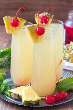 two glasses filled with pineapple and cherries on a plate next to other drinks