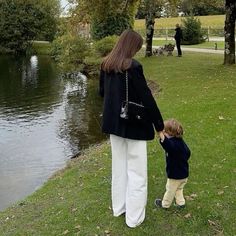 a woman holding the hand of a small child near a pond with trees and people in the background