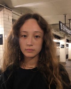 a young woman with long hair standing in a subway station looking at the camera and to her left