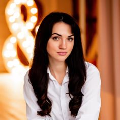 a woman with long dark hair wearing a white shirt and sitting in front of a bed