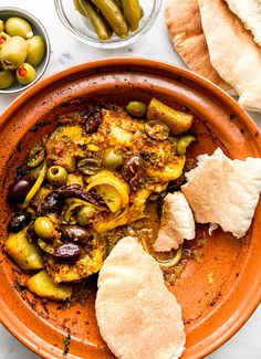 an orange bowl filled with food next to bread and olives