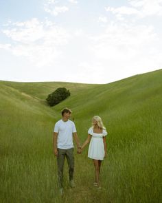 a young man and woman holding hands walking through tall grass in the middle of a field