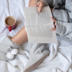 a person sitting on a bed reading a book and holding a cup of coffee in their hands