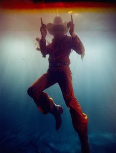 a man in cowboy hat and orange pants under water with his hands up to the air