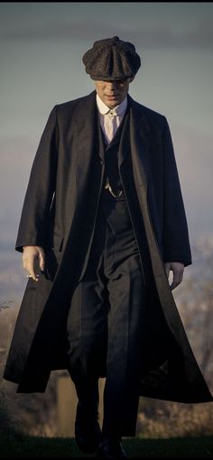 a man in a black suit and hat walking down a hill with his hands in his pockets