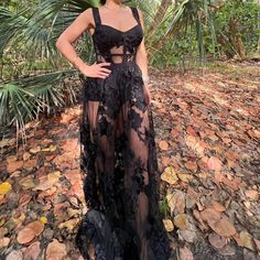 a woman standing in the woods wearing a black dress with sheer fabric and flowers on it