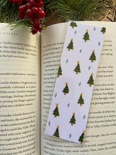 an open book with christmas trees on it next to holly branches and red berries in the background