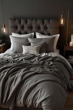 a bed with grey linens and pillows in a dark colored room, lit by two lamps