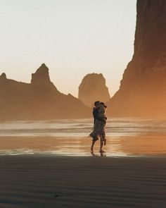 two people are hugging on the beach near some mountains and water at sunset or sunrise